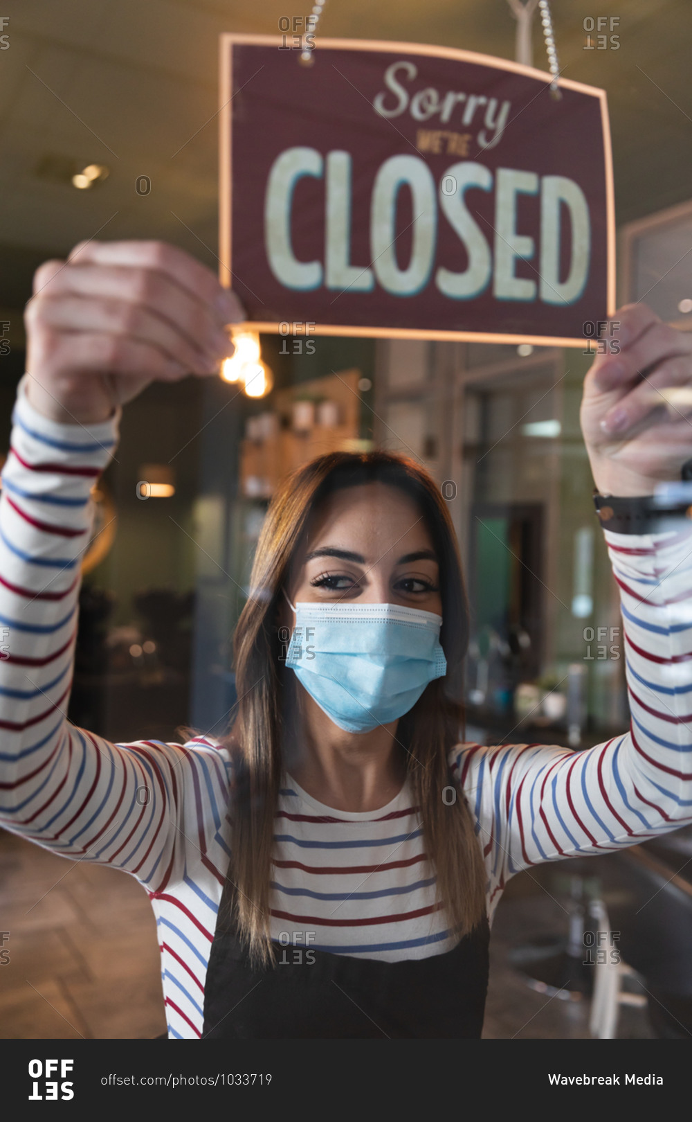 Caucasian female hairdresser working in hair salon wearing face mask, holding Sorry, We Are Closed sign. Health and hygiene in workplace during Coronavirus Covid 19 pandemic.