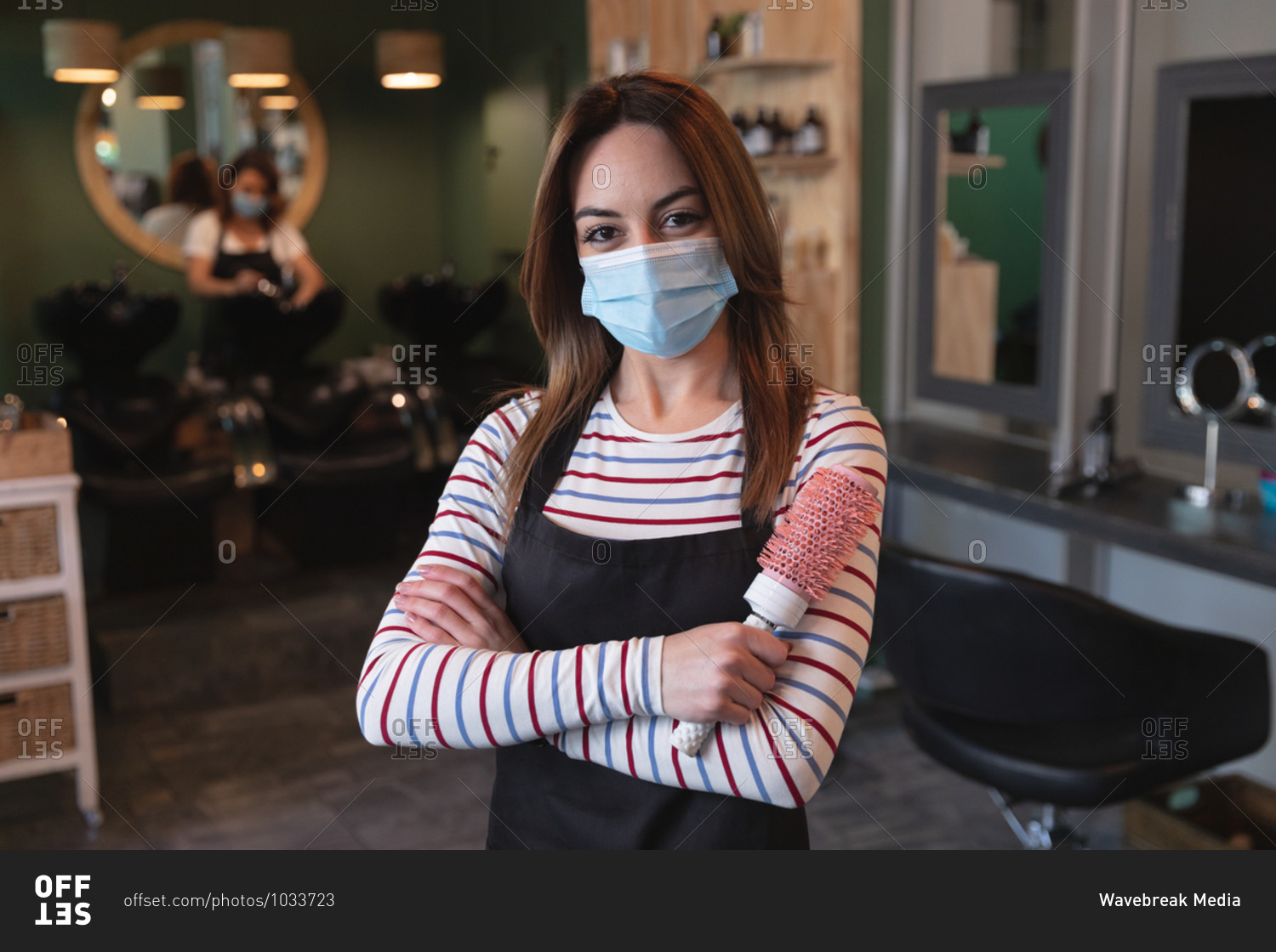 Portrait of a Caucasian female hairdresser working in hair salon wearing face mask, posing for a photo, holding a hairbrush. Health and hygiene in workplace during Coronavirus Covid 19 pandemic.