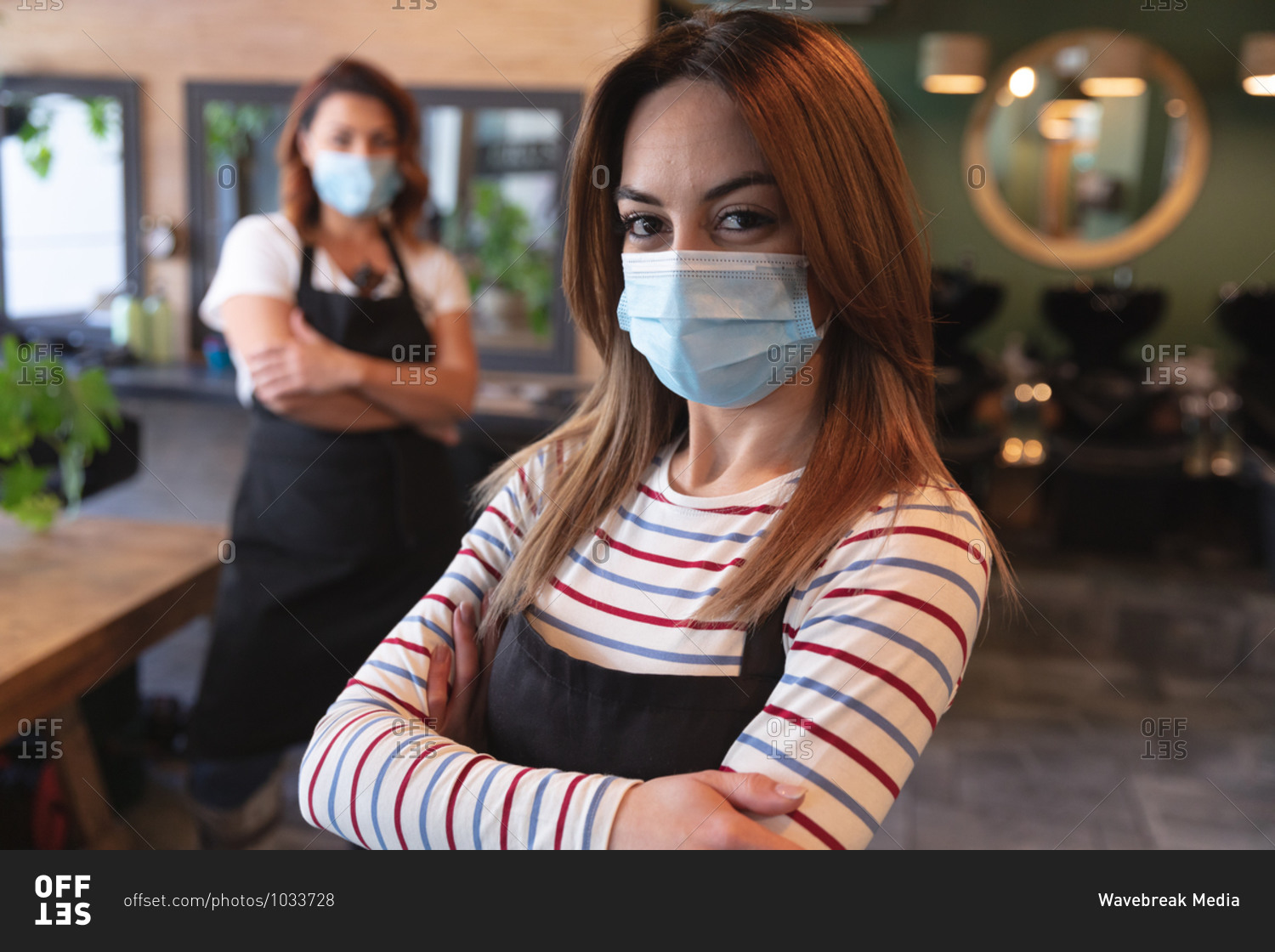 Portrait of Caucasian female hairdresser working in hair salon wearing face mask, posing for a picture with her arms crossed. Health and hygiene in workplace during Coronavirus Covid 19 pandemic.