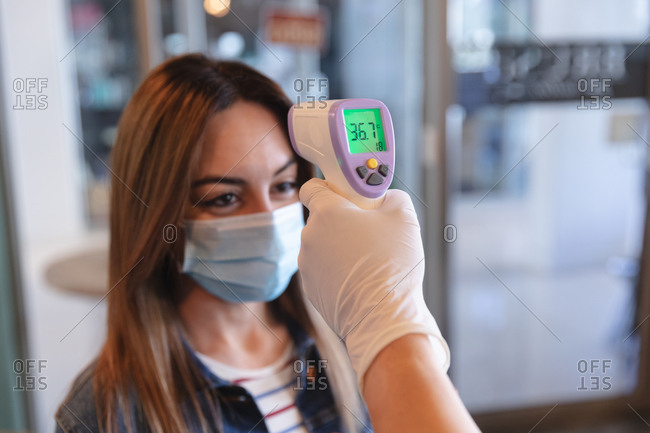 Caucasian female hairdresser working in hair salon, measuring temperature of a female Caucasian customer in face mask. Health and hygiene in workplace during Coronavirus Covid 19 pandemic.