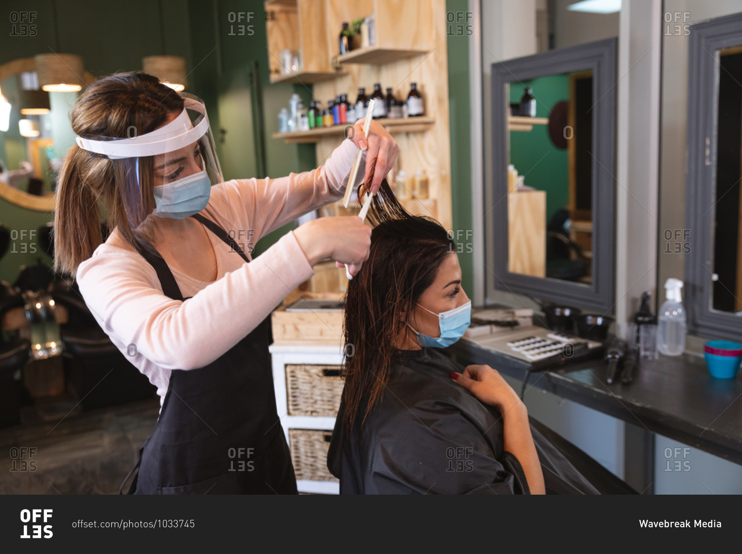 Caucasian female hairdresser working in hair salon wearing face mask, combing hair of female Caucasian customer in face mask. Health and hygiene in workplace during Coronavirus Covid 19 pandemic.