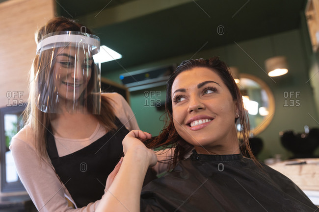 Caucasian female hairdresser working in hair salon wearing face cover, combing hair of female Caucasian customer smiling. Health and hygiene in workplace during Coronavirus Covid 19 pandemic.