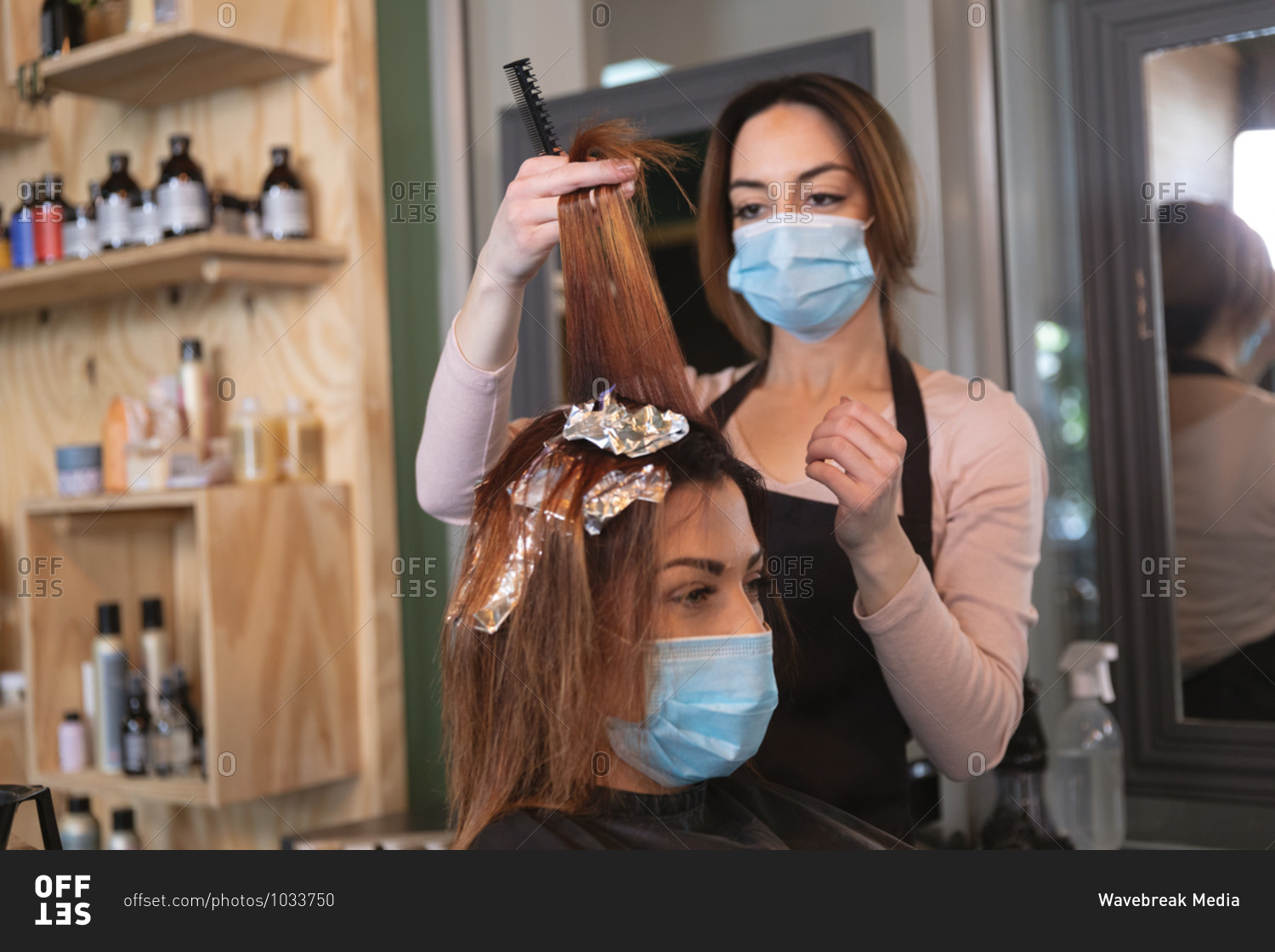 Caucasian female hairdresser working in hair salon wearing face mask, dying hair of female Caucasian customer in face mask. Health and hygiene in workplace during Coronavirus Covid 19 pandemic.