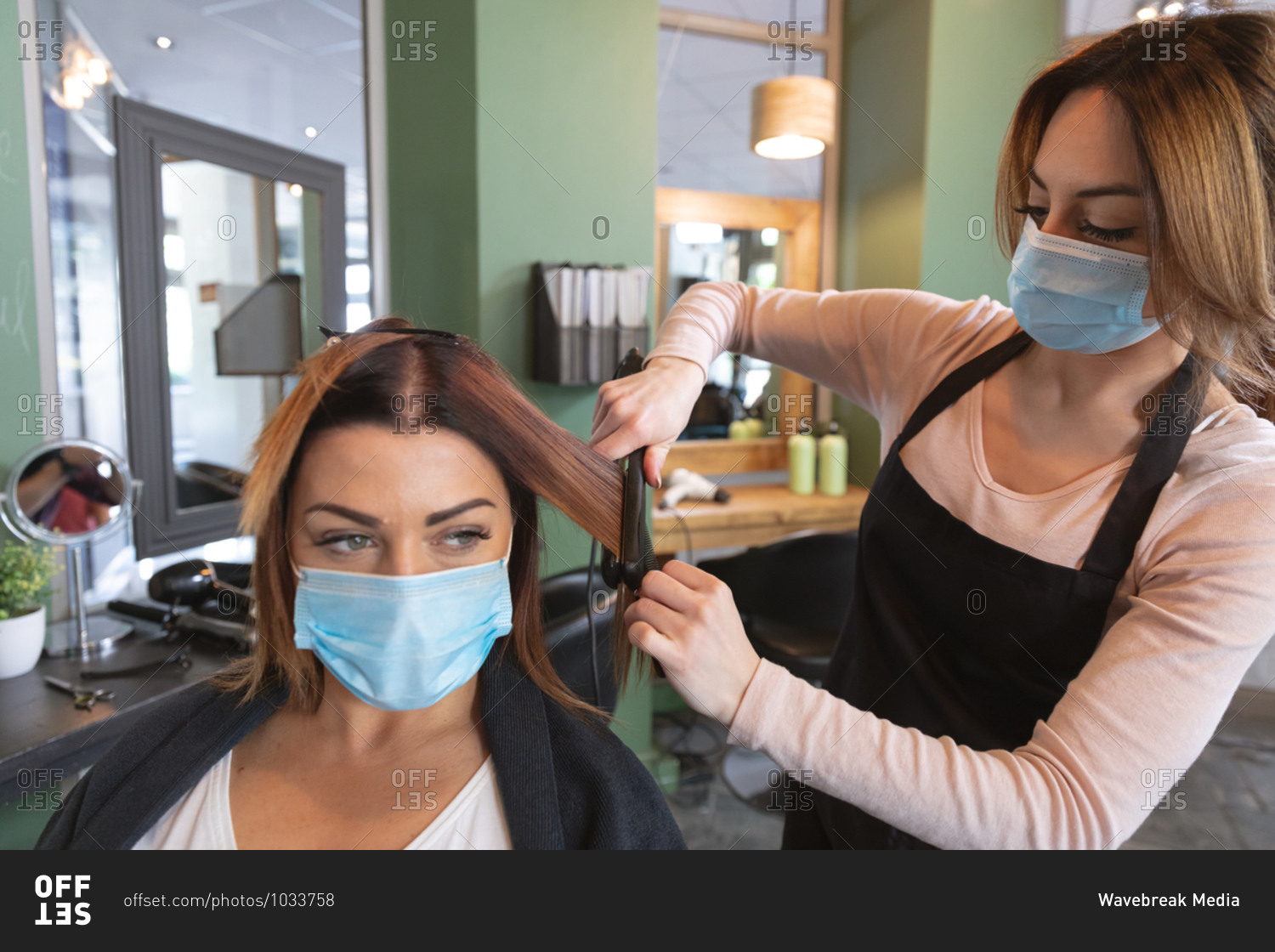 Caucasian female hairdresser working in hair salon wearing face mask straightening hair of female Caucasian customer in face mask. Health and hygiene in workplace during Coronavirus Covid 19 pandemic.