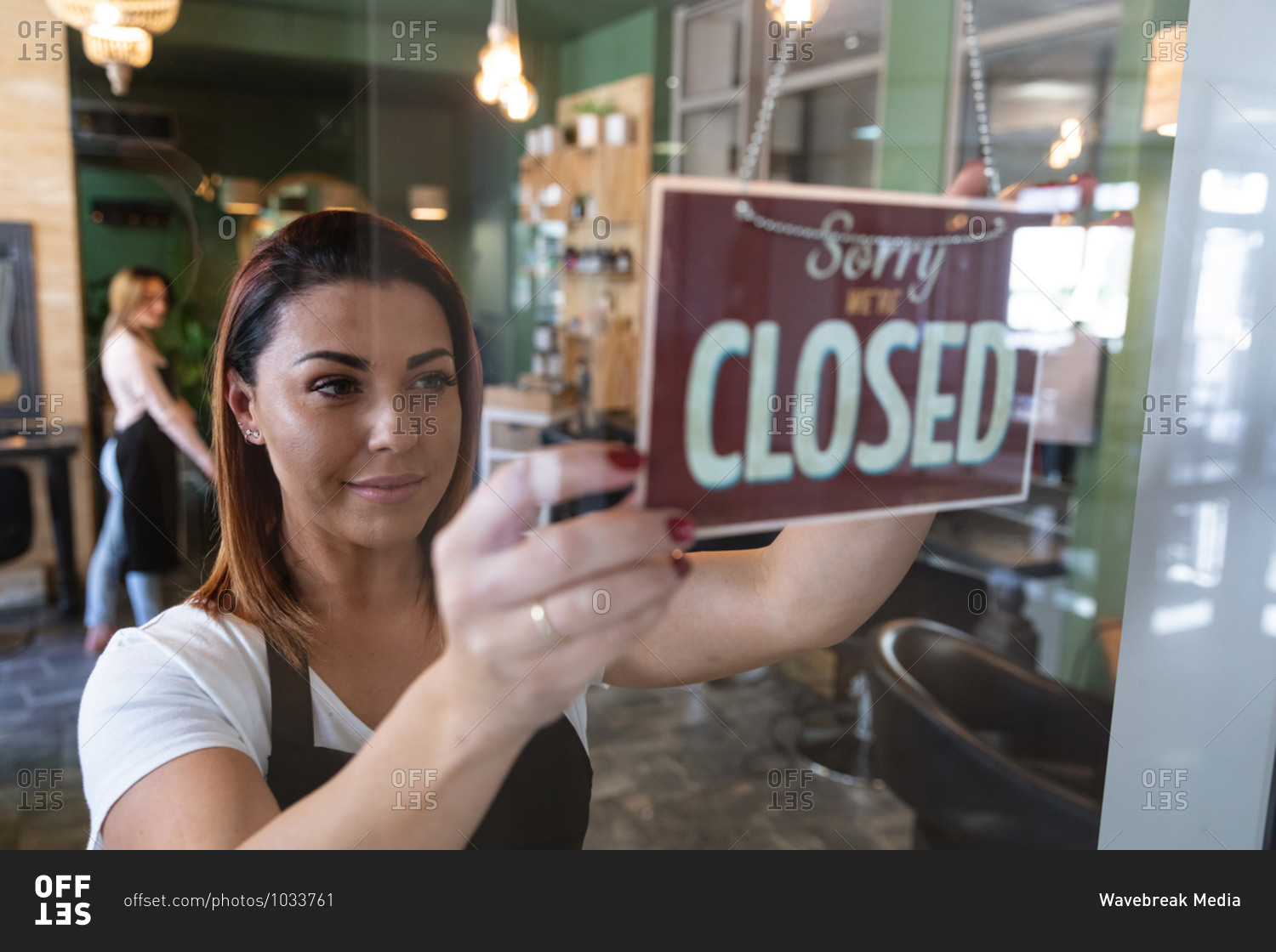 Caucasian female hairdresser working in hair salon smiling, fixing a sign saying Sorry, We Are Closed. Health and hygiene in workplace during Coronavirus Covid 19 pandemic.