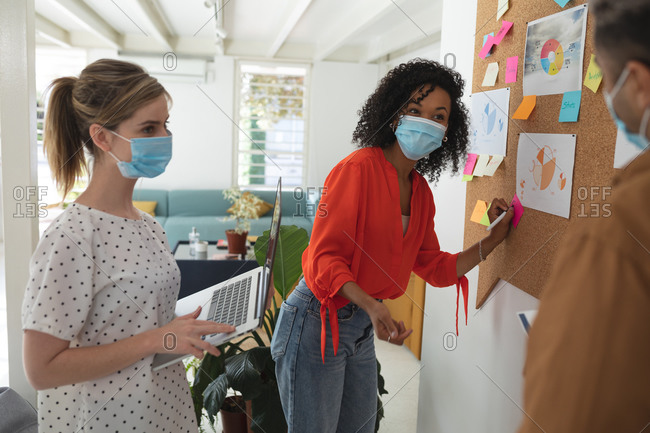 Multi ethnic group of male and female business creatives stand brainstorming in modern office wearing face masks. Health and hygiene in the workplace during Coronavirus Covid 19 pandemic.