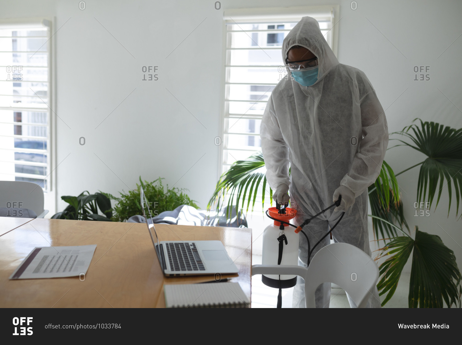 Mixed race male cleaner wearing anti-contamination overalls spraying disinfecting an office. Health and hygiene in the workplace during Coronavirus Covid 19 pandemic.