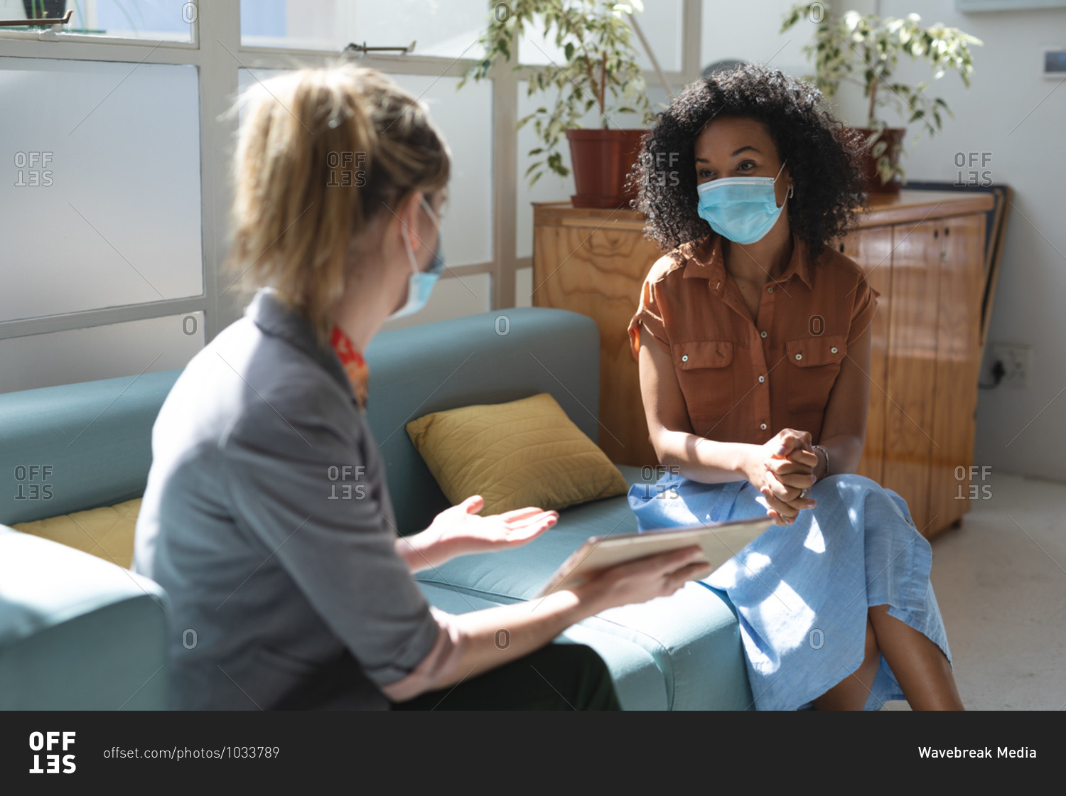 Mixed race and Caucasian female business creatives wearing face masks and distancing on sofa, talking and using tablet in office. Health and hygiene in workplace during Coronavirus Covid 19 pandemic.