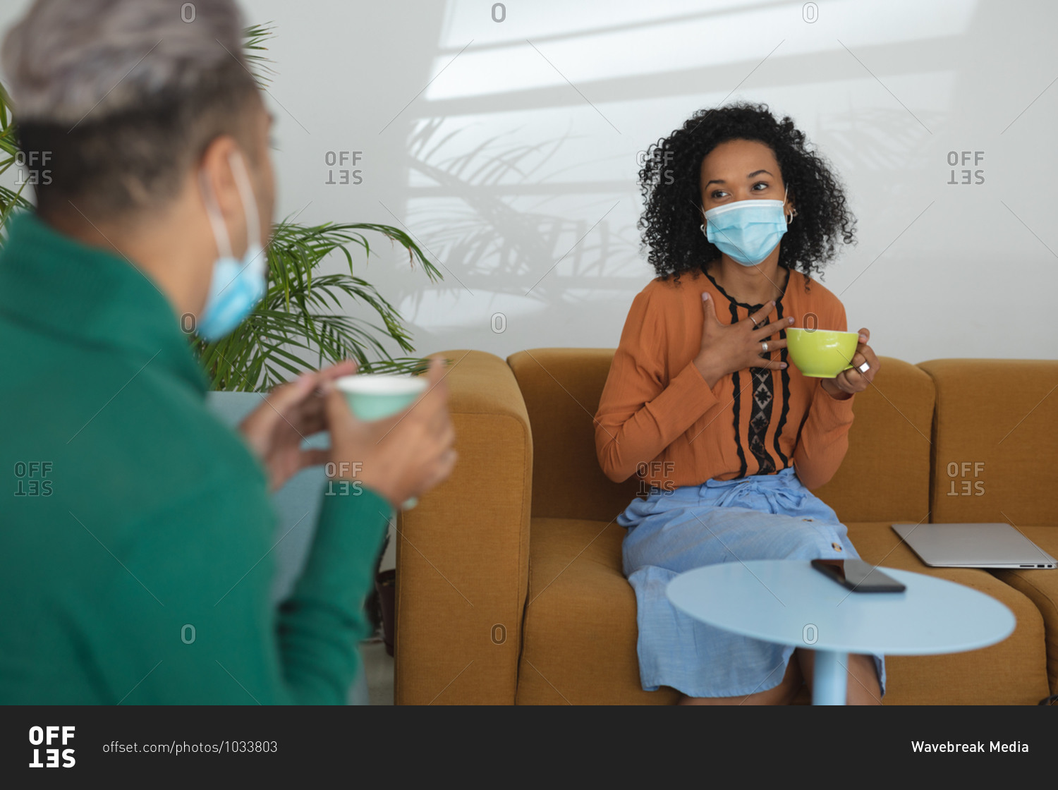 Mixed race male and female business creatives wearing face masks and distancing holding coffees and talking in office lounge. Health and hygiene in workplace during Coronavirus Covid 19 pandemic.