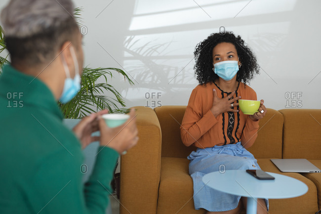 Mixed race male and female business creatives wearing face masks and distancing holding coffees and talking in office lounge. Health and hygiene in workplace during Coronavirus Covid 19 pandemic.