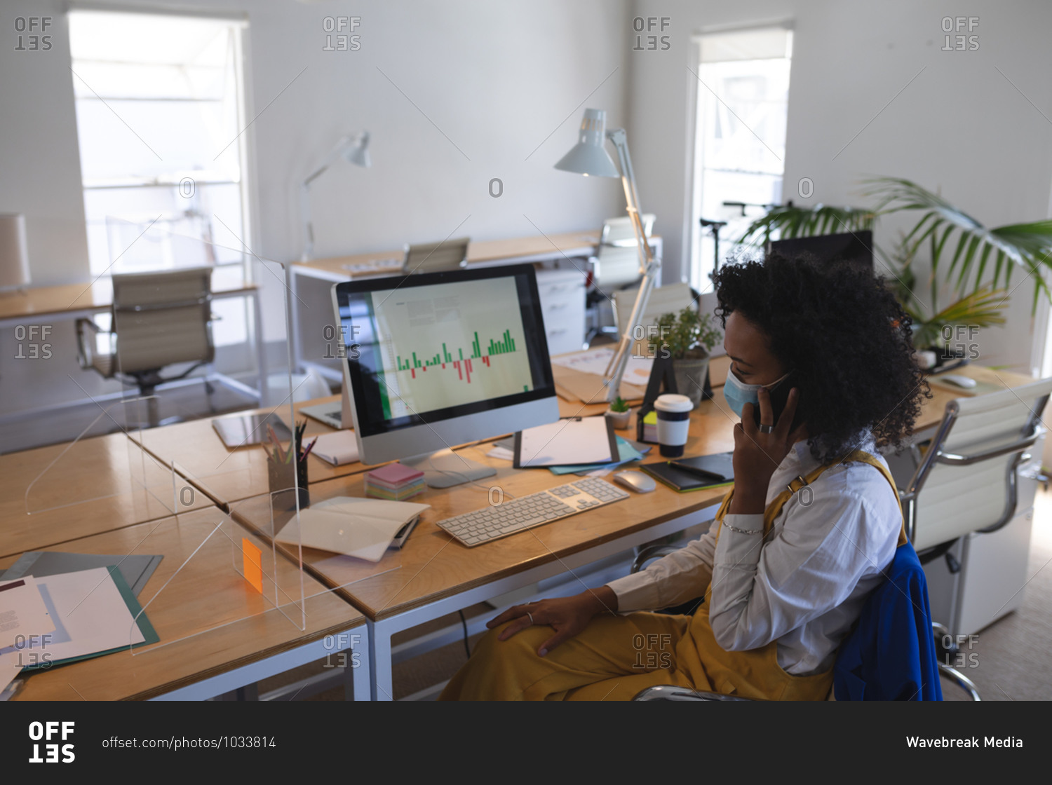 Mixed race woman sitting at desk in a modern office wearing a face mask, talking on smartphone and using computer. Health and hygiene in the workplace during Coronavirus Covid 19 pandemic.