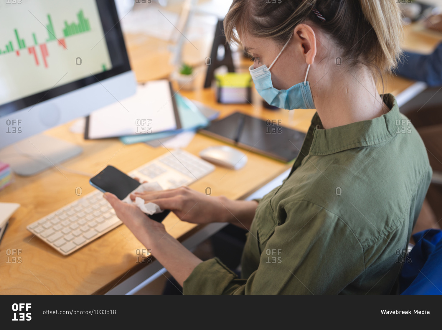 Caucasian female business creative sitting at desk in an office wearing face mask disinfecting her smartphone. Health and hygiene in workplace during Coronavirus Covid 19 pandemic.
