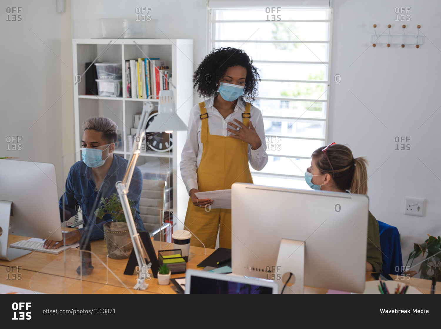 Mixed race and Caucasian female creative business colleague talking in office wearing face masks, male colleague in background. Health and hygiene in workplace during Coronavirus Covid 19 pandemic.