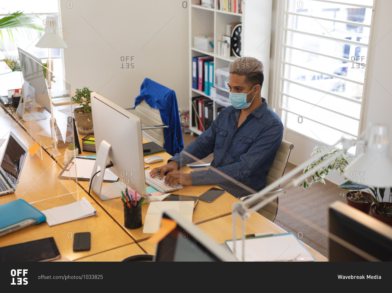 Mixed race male creative sitting at desk in a modern office, wearing a face mask and using a computer. Health and hygiene in the workplace during Coronavirus Covid 19 pandemic.