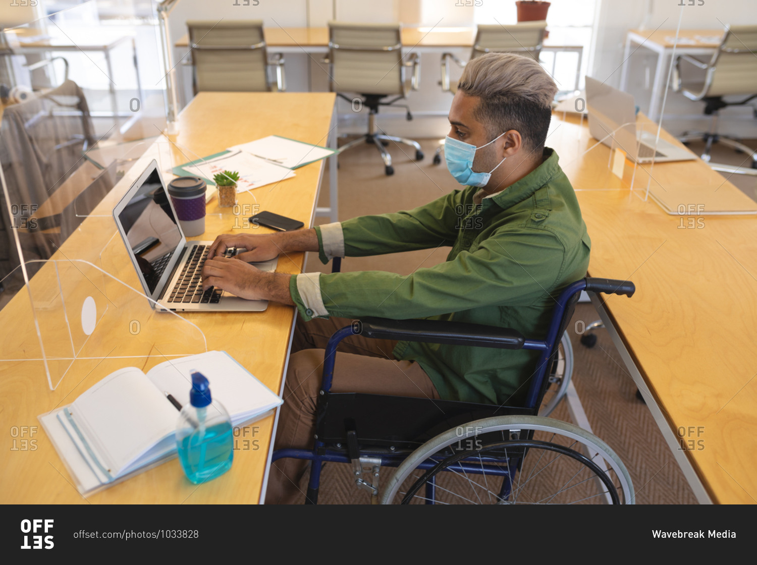 Mixed race male creative sitting in a wheelchair at desk in an office, wearing face mask, using a laptop computer. Health and hygiene in workplace during Coronavirus Covid 19 pandemic.