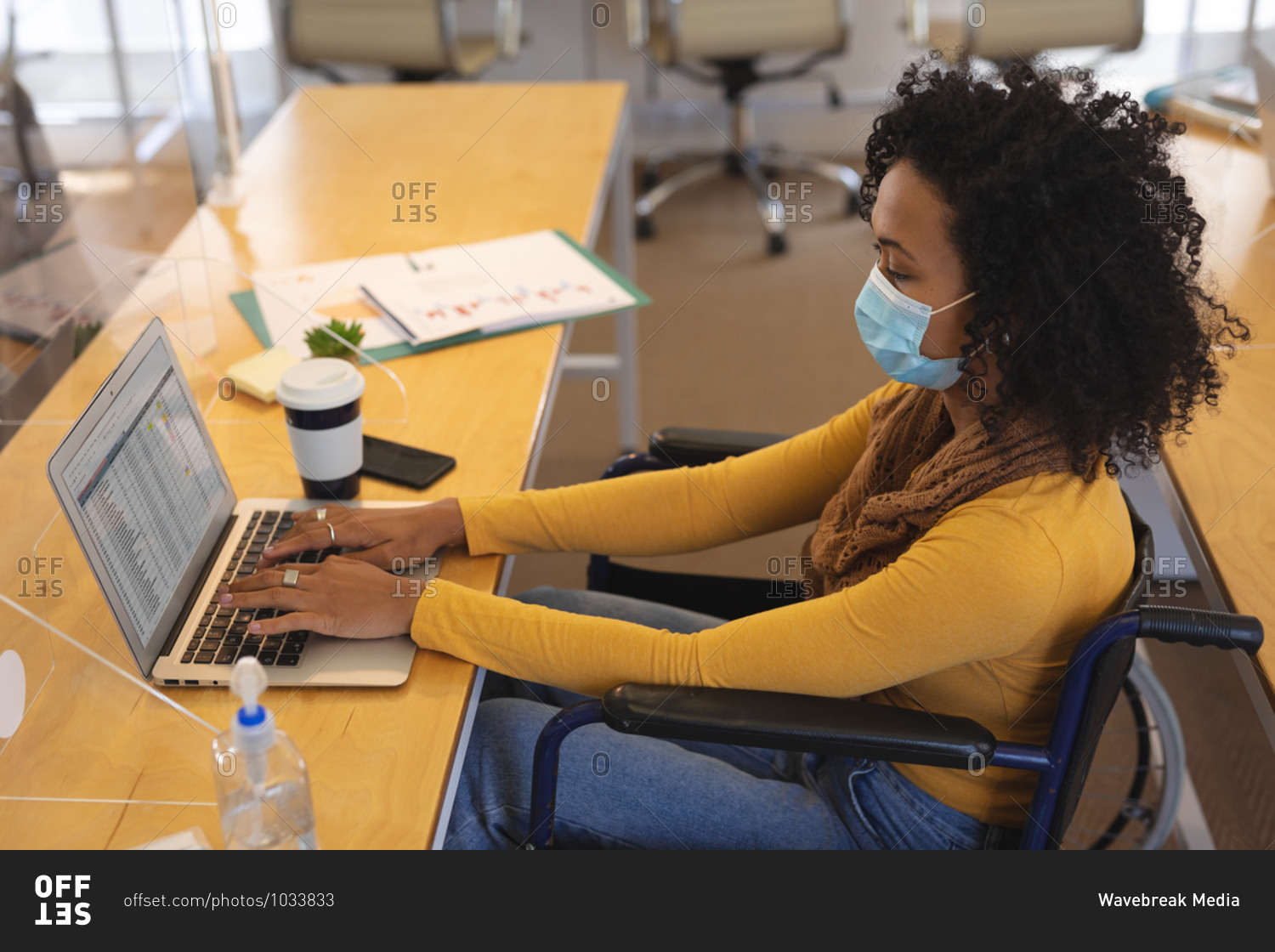 Mixed race female creative sitting in a wheelchair at desk in an office, wearing face mask, using a laptop computer. Health and hygiene in workplace during Coronavirus Covid 19 pandemic.