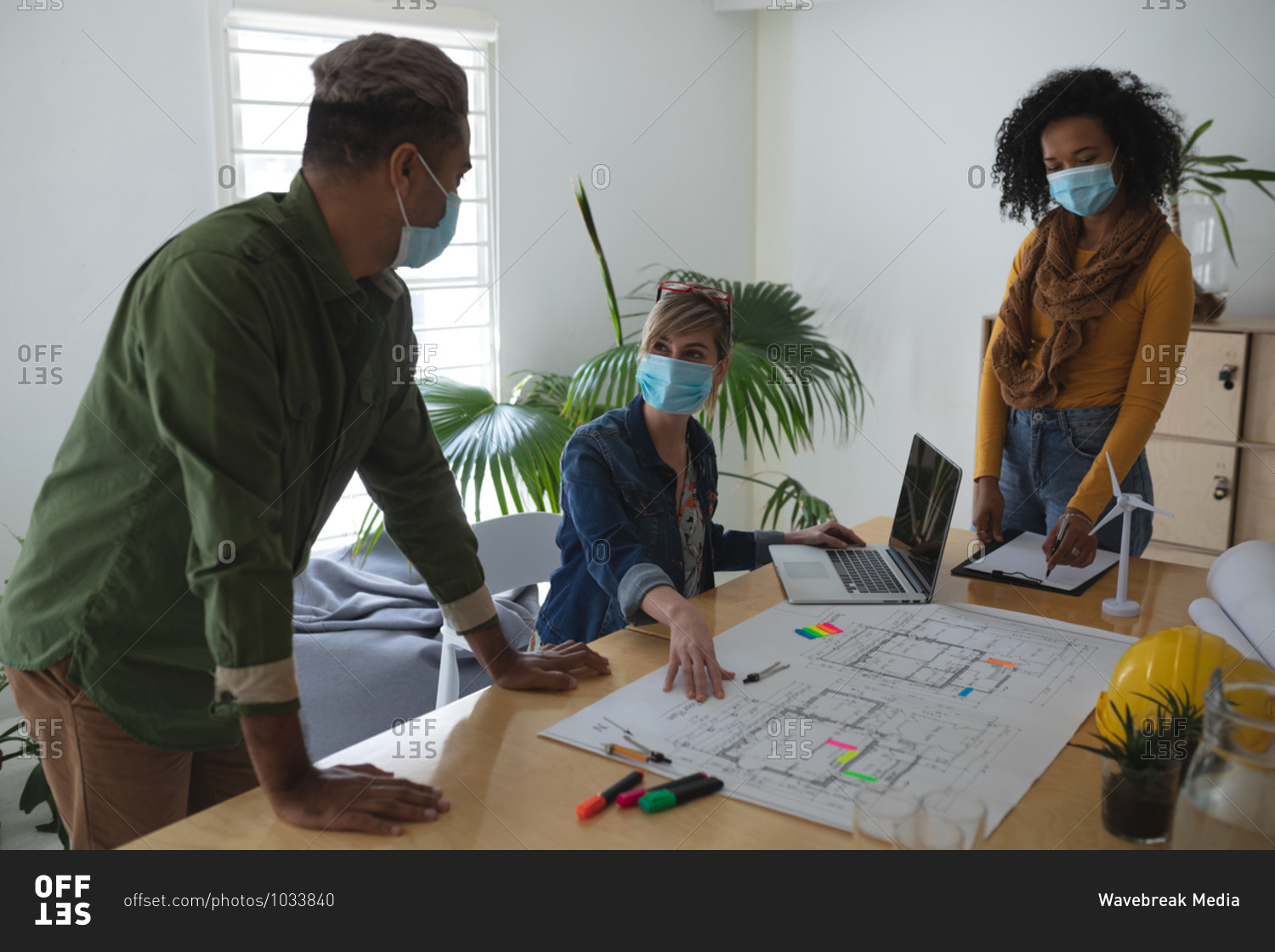 Multi ethnic group of male and female architects in office wearing face masks, discussing over architectural drawing. Health and hygiene in workplace during Coronavirus Covid 19 pandemic.
