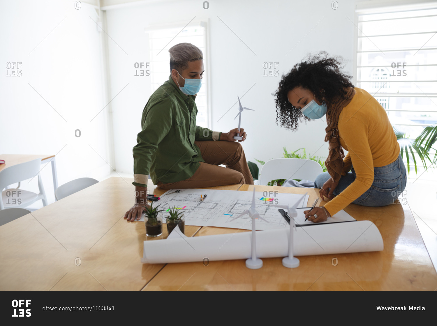 Mixed race male and female architects in office wearing face masks, discussing over architectural drawing. Health and hygiene in workplace during Coronavirus Covid 19 pandemic.