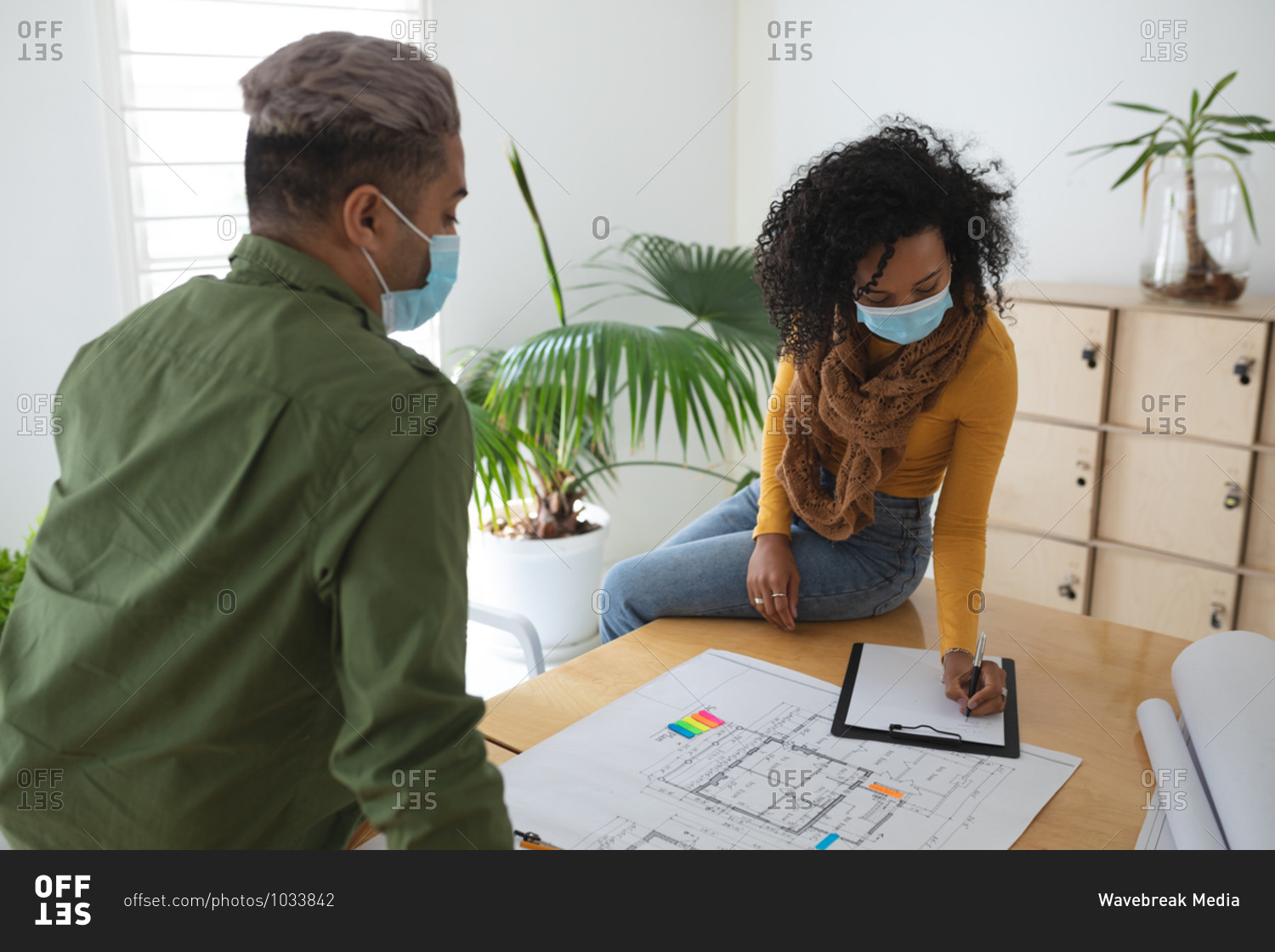 Mixed race male and female architects in office wearing face masks, discussing over architectural drawing. Health and hygiene in workplace during Coronavirus Covid 19 pandemic.