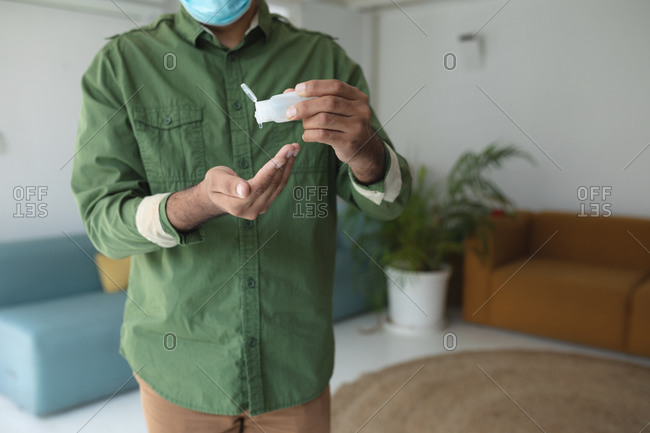 Mixed race man in modern office wearing face mask, disinfecting hands with hand sanitizer. Health and hygiene in workplace during Coronavirus Covid 19 pandemic.