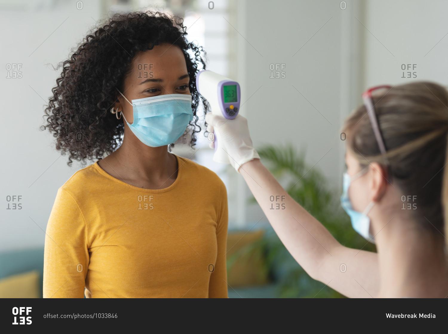 Caucasian woman using digital thermometer taking temperature of mixed race woman arriving at work wearing face mask. Health and hygiene in workplace during Coronavirus Covid 19 pandemic.