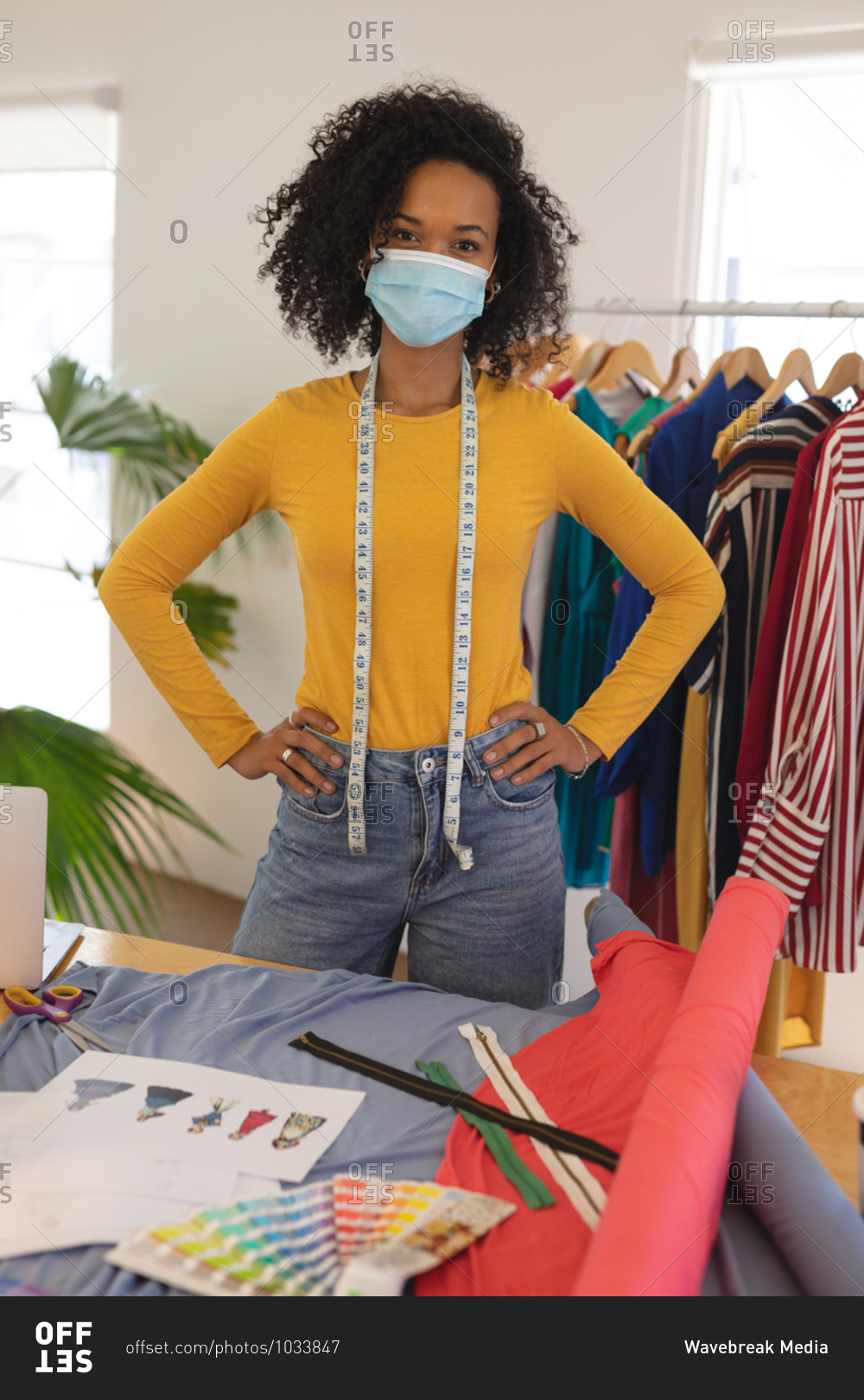 Portrait of mixed race female fashion designer in studio wearing face mask and tape measure around her neck, looking at camera. Health and hygiene in workplace during Coronavirus Covid 19 pandemic.