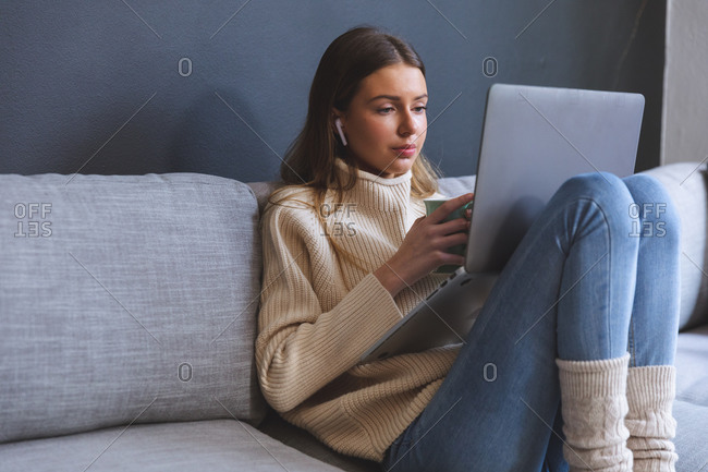 Caucasian woman spending time at home, sitting on sofa in sitting room using laptop computer with earphones, holding mug. Social distancing during Covid 19 Coronavirus quarantine lockdown.