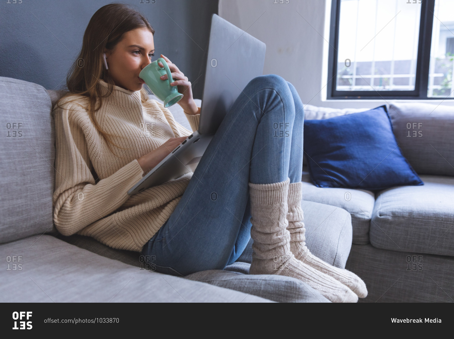 Caucasian woman spending time at home, sitting on sofa in sitting room using laptop computer with earphones, holding mug, drinking. Social distancing during Covid 19 Coronavirus quarantine lockdown.
