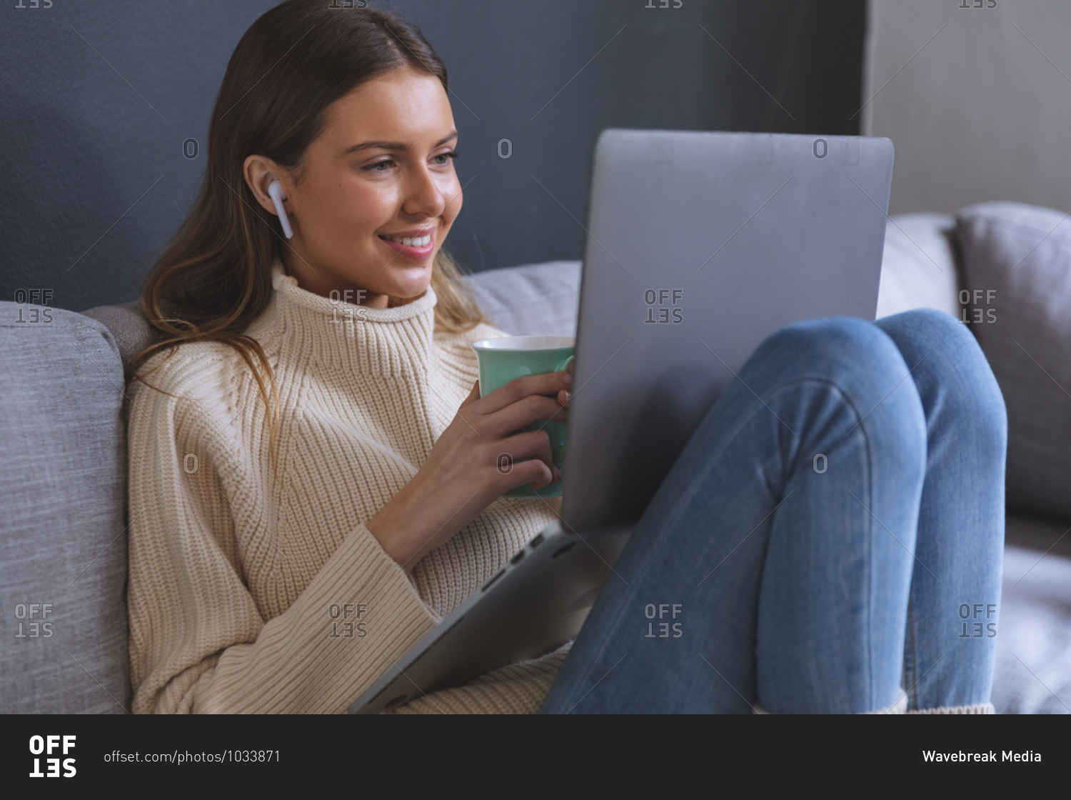 Smiling Caucasian woman spending time at home, sitting on sofa in sitting room using laptop computer with earphones, holding mug. Social distancing during Covid 19 Coronavirus quarantine lockdown.