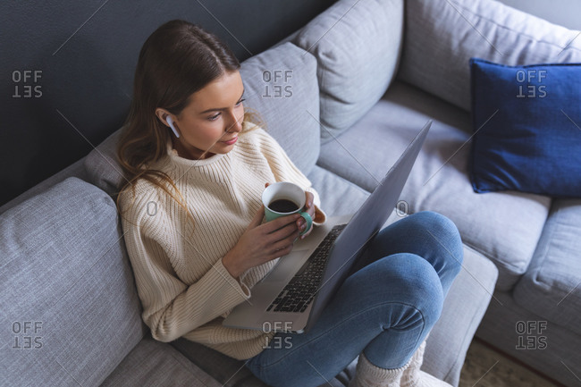 Caucasian woman spending time at home, sitting on sofa in sitting room using laptop computer with earphones, holding mug. Social distancing during Covid 19 Coronavirus quarantine lockdown.