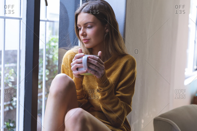 Caucasian woman spending time at home, sitting on windowsill in living room, holding green mug. Social distancing during Covid 19 Coronavirus quarantine lockdown.