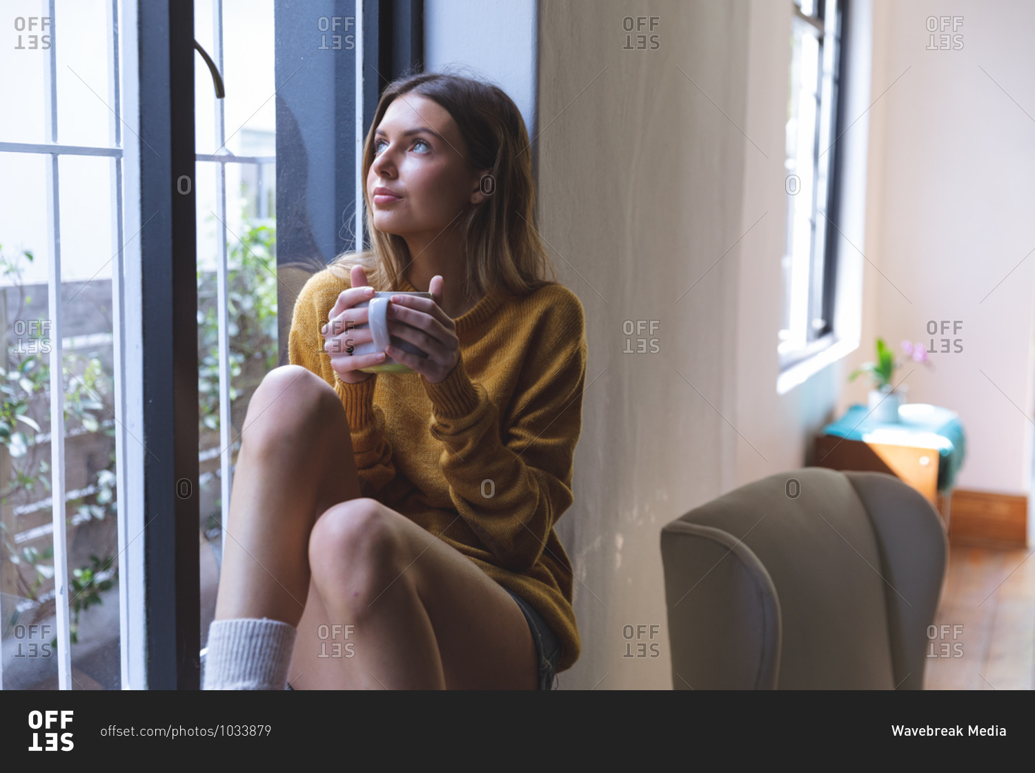 Caucasian woman spending time at home, sitting on windowsill in living room, holding green mug looking out of window. Social distancing during Covid 19 Coronavirus quarantine lockdown.