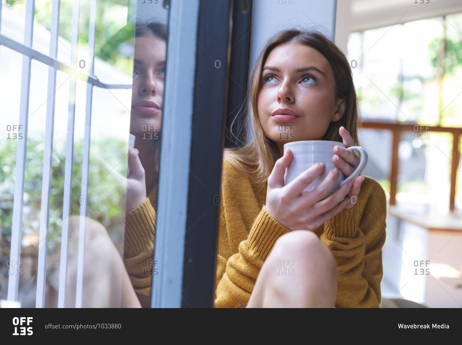 Caucasian woman spending time at home, sitting by window, holding green mug looking out of window. Social distancing during Covid 19 Coronavirus quarantine lockdown.