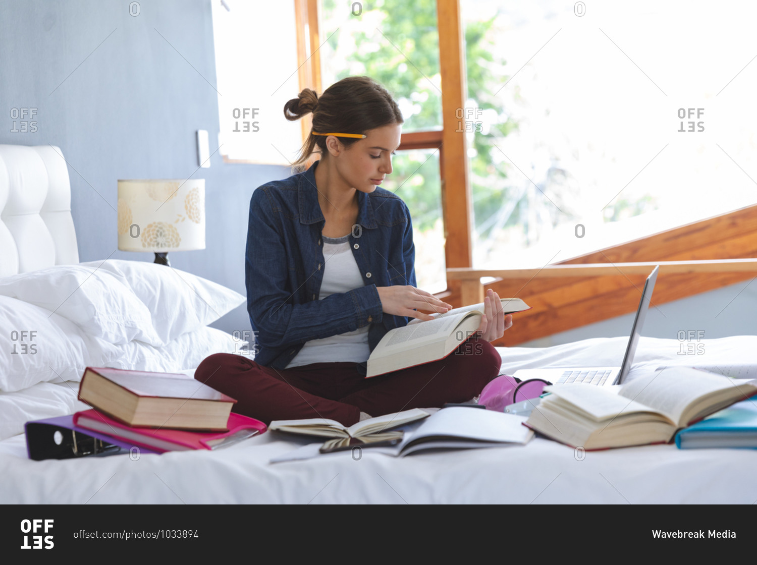 Caucasian woman spending time at home, sitting on bed in bedroom, studying from home, holding and reading book. Social distancing during Covid 19 Coronavirus quarantine lockdown.