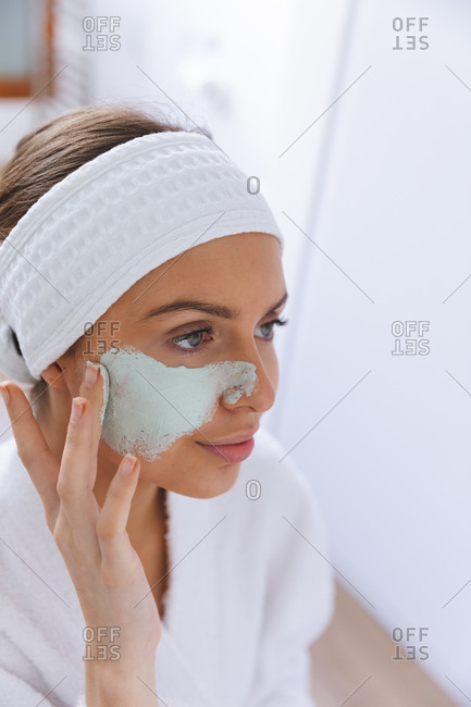 Caucasian woman spending time at home, standing in bathroom, looking in mirror applying face mask. Social distancing during Covid 19 Coronavirus quarantine lockdown.