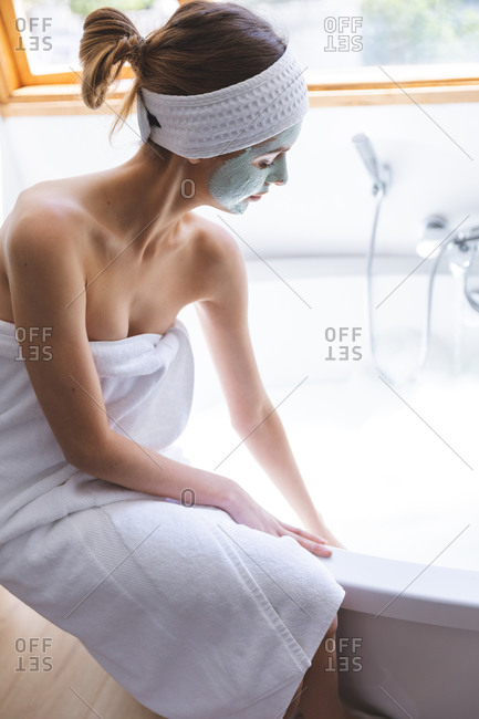 Caucasian woman spending time at home, in bathroom with face mask on, running bath sitting on edge of bathtub. Social distancing during Covid 19 Coronavirus quarantine lockdown.