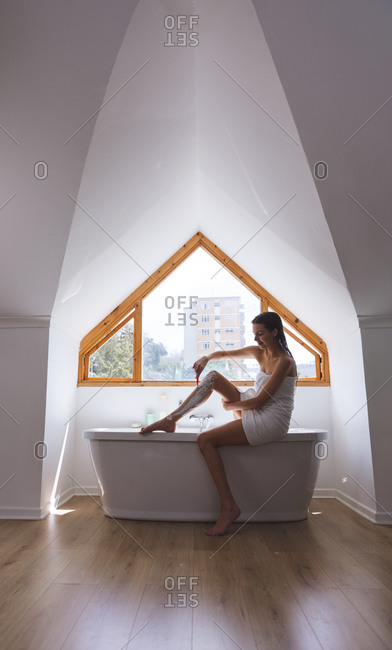 Caucasian woman spending time at home, in bathroom, sitting on bathtub edge, shaving legs. Social distancing during Covid 19 Coronavirus quarantine lockdown.