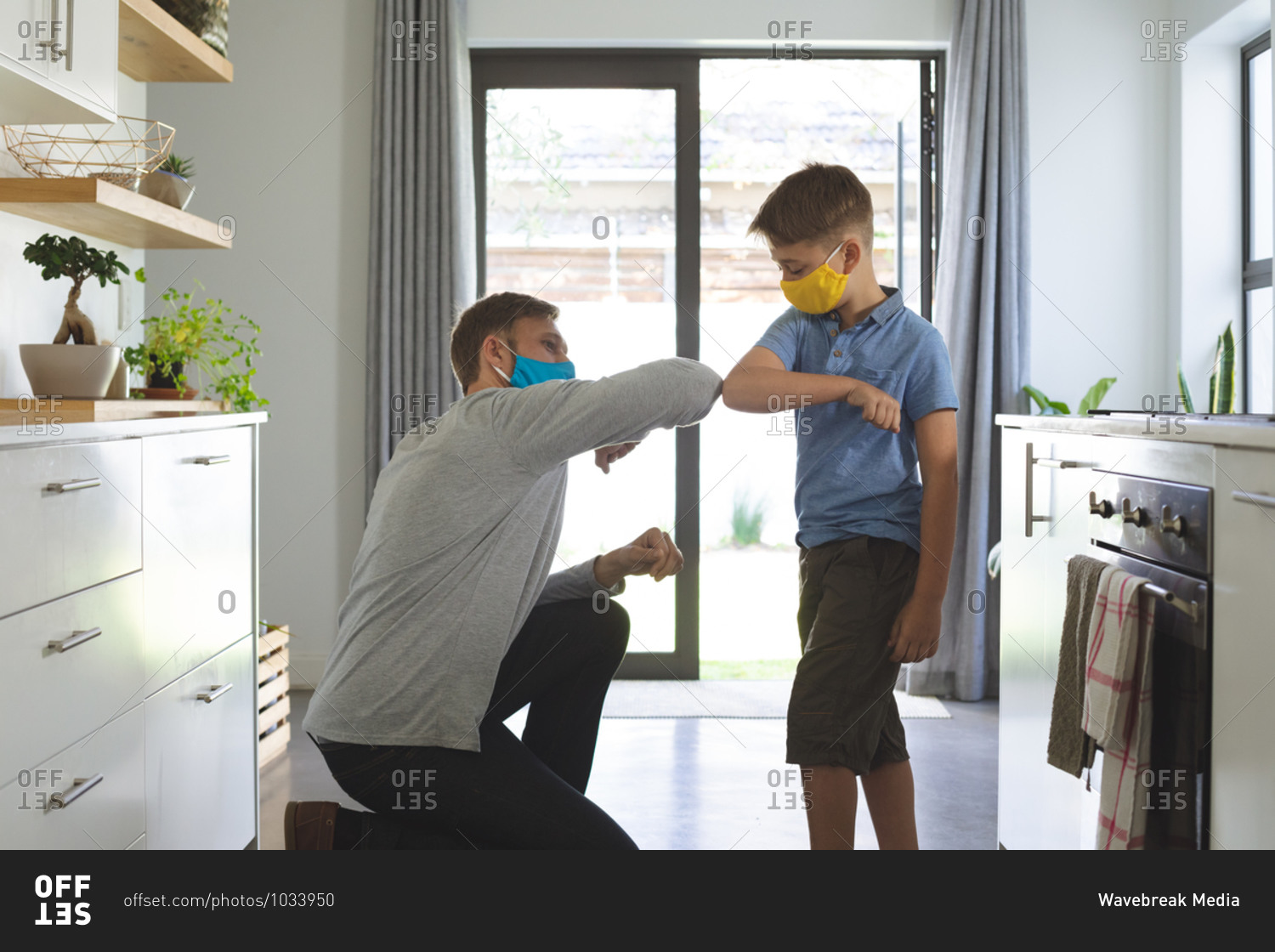 Caucasian man at home with his son, in kitchen, wearing face masks, greeting touching elbows. Social distancing during Covid 19 Coronavirus quarantine lockdown.