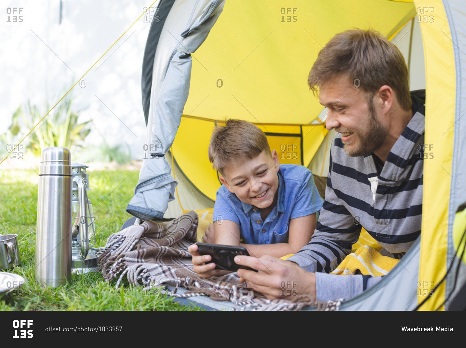 Caucasian man spending time with his son together, camping in garden, lying in tent using smartphone, smiling. Social distancing during Covid 19 Coronavirus quarantine lockdown.