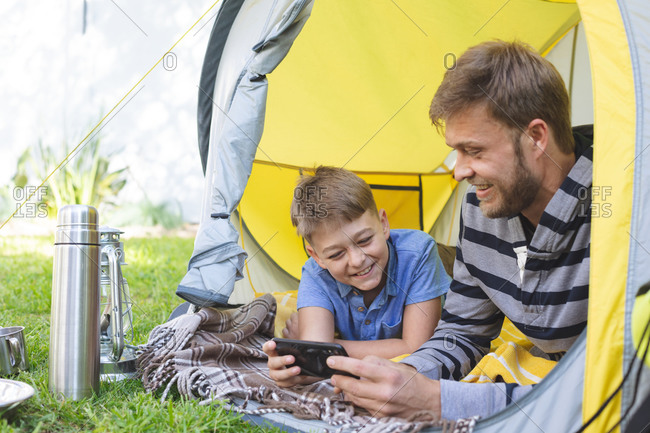 Caucasian man spending time with his son together, camping in garden, lying in tent using smartphone, smiling. Social distancing during Covid 19 Coronavirus quarantine lockdown.