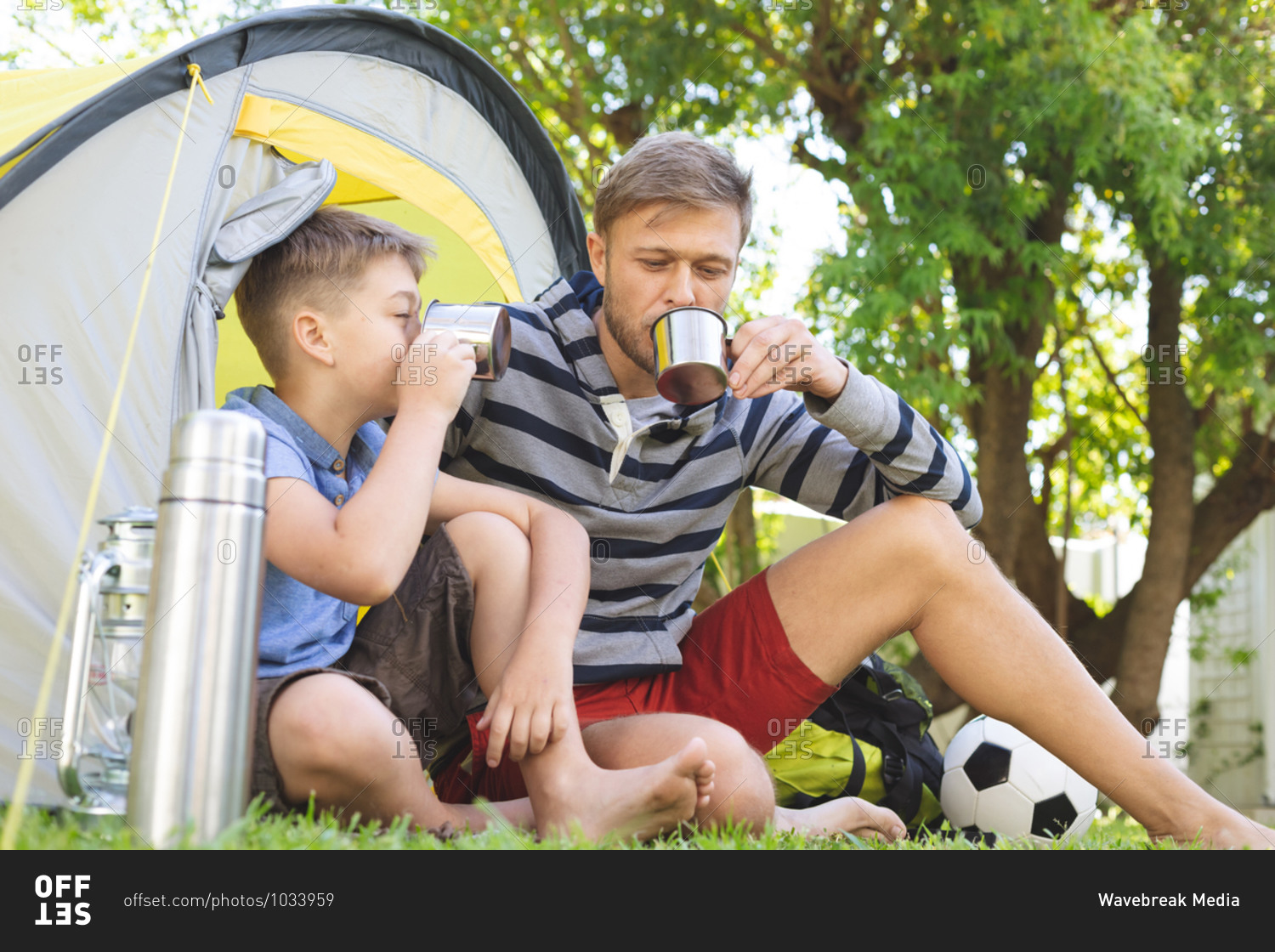 Caucasian man spending time with his son together, camping in garden, sitting in tent, drinking tea. Social distancing during Covid 19 Coronavirus quarantine lockdown.
