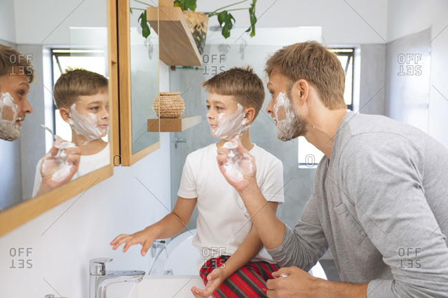 Caucasian man at home with his son together, in bathroom, shaving with shaving cream on faces, looking at mirror. Social distancing during Covid 19 Coronavirus quarantine lockdown.