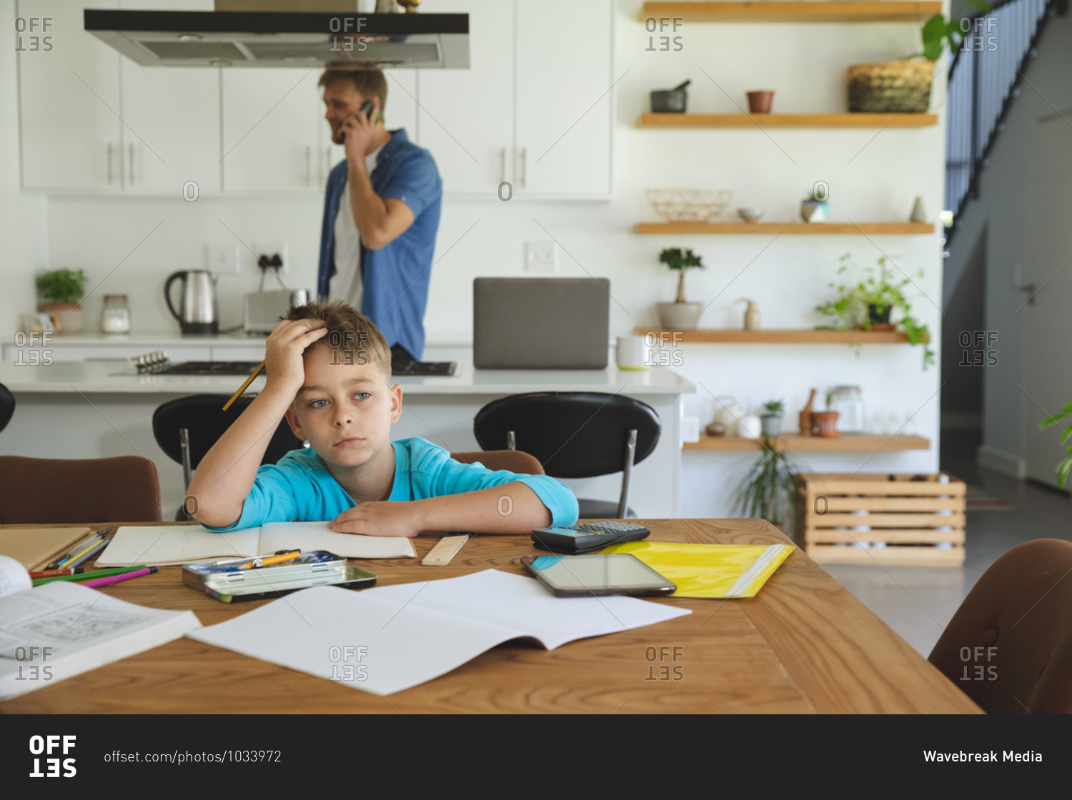 Caucasian man at home with his son together, in kitchen, boy doing homework at table, thinking. Social distancing during Covid 19 Coronavirus quarantine lockdown.