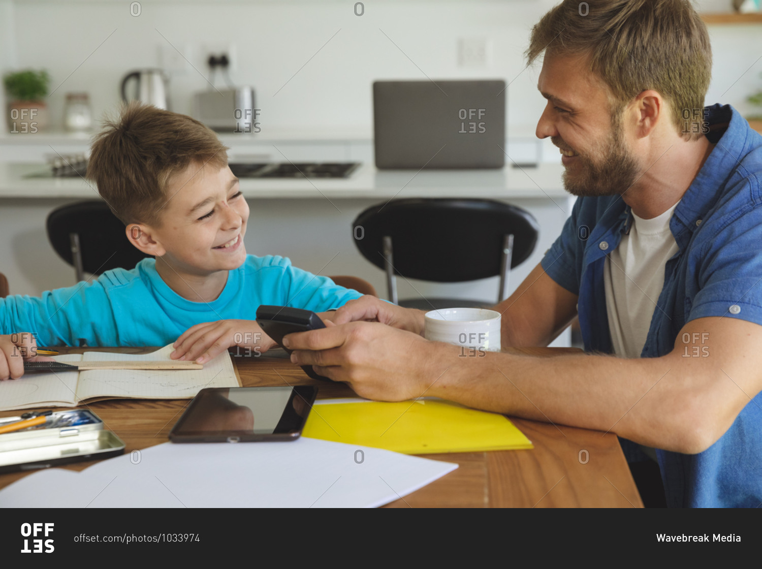 Caucasian man at home with his son together, in kitchen, father helping the boy doing homework at table. Social distancing during Covid 19 Coronavirus quarantine lockdown.
