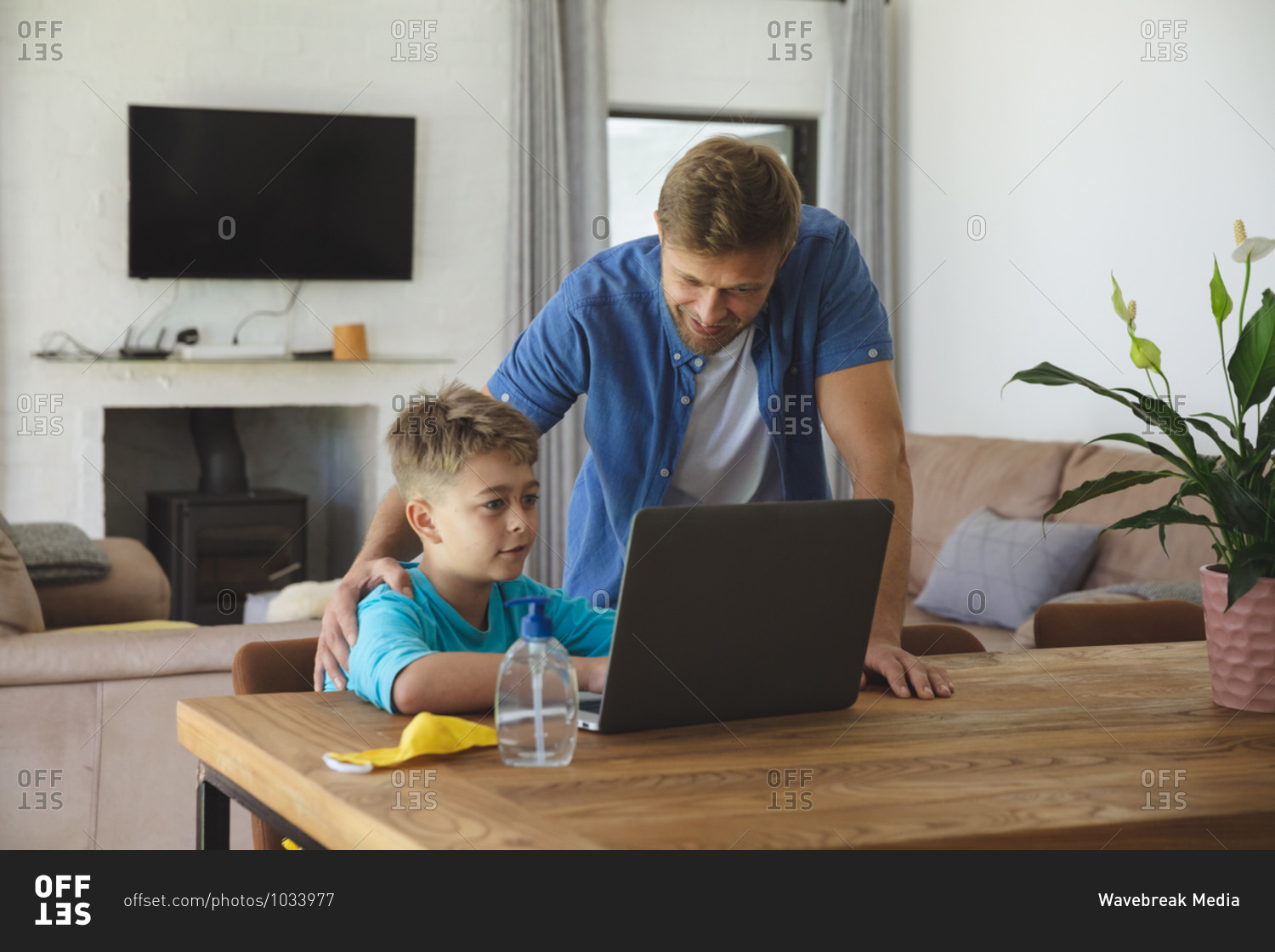 Caucasian man at home with his son together, boy sitting at table, using laptop computer, father standing next to him. Social distancing during Covid 19 Coronavirus quarantine lockdown.