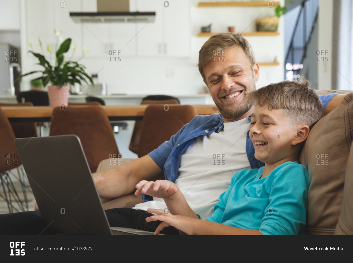 Caucasian man at home with his son together, sitting on sofa in living room, using laptop computer, smiling. Social distancing during Covid 19 Coronavirus quarantine lockdown.