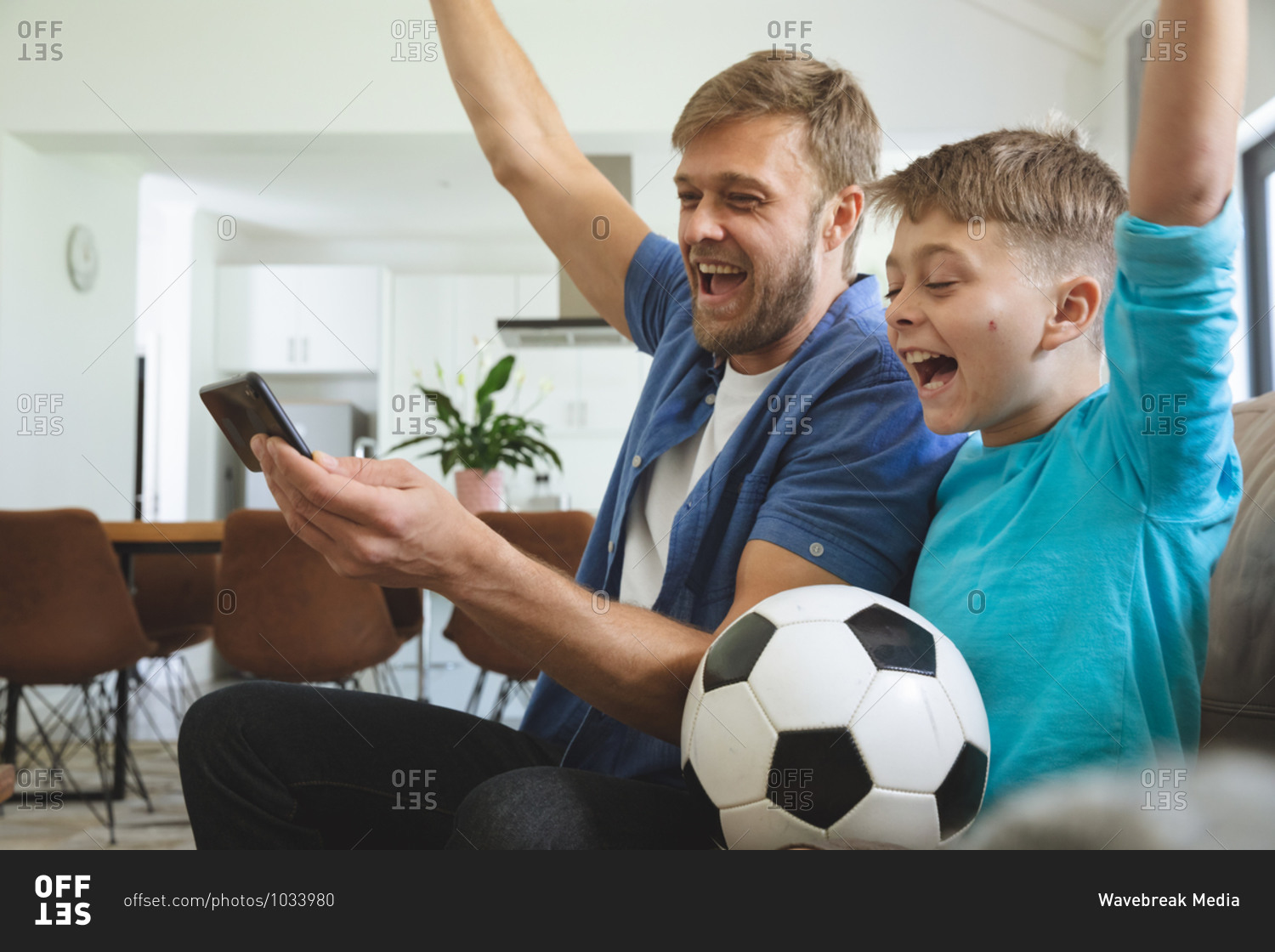 Caucasian man at home with his son together, sitting on sofa in living room, watching sports football game on smartphone, cheering. Social distancing during Covid 19 Coronavirus quarantine lockdown.