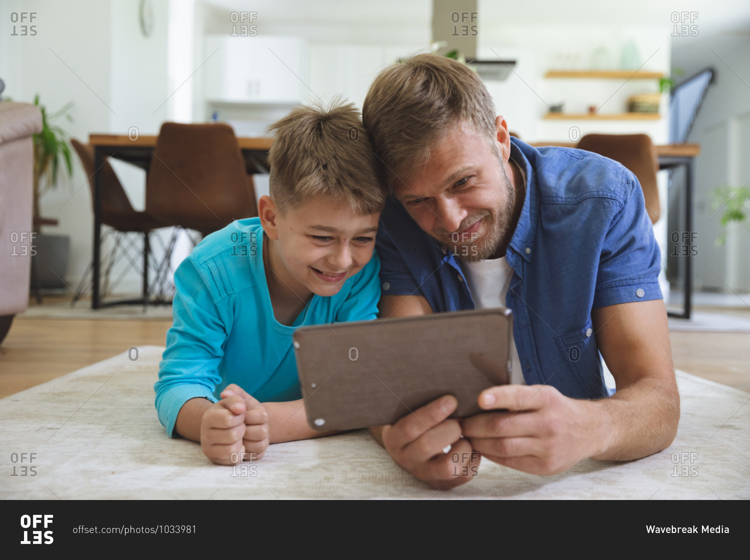 Caucasian man at home with his son together, lying on rug in living room, using digital tablet, smiling. Social distancing during Covid 19 Coronavirus quarantine lockdown.