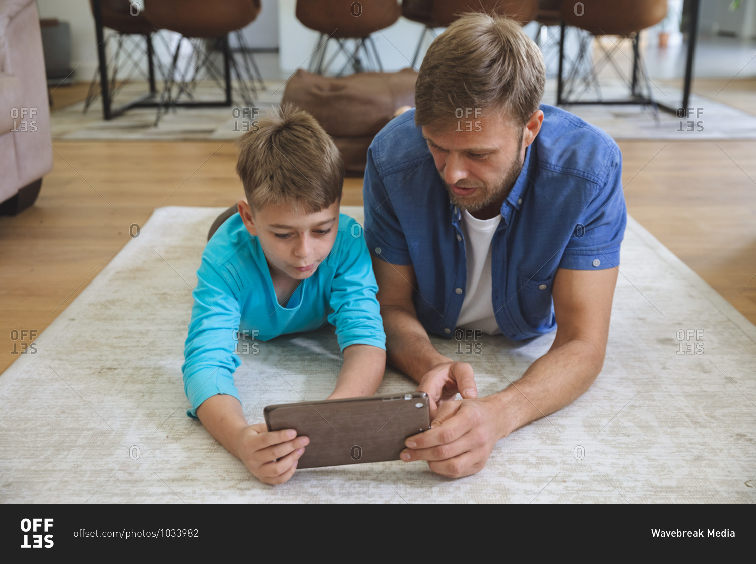 Caucasian man at home with his son together, lying on rug in living room, using digital tablet. Social distancing during Covid 19 Coronavirus quarantine lockdown.