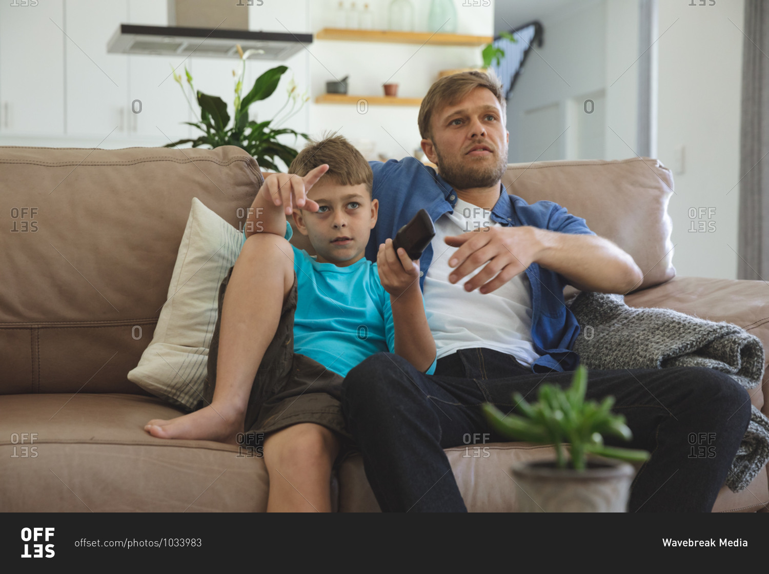 Caucasian man at home with his son together, sitting on sofa in living room, watching TV. Social distancing during Covid 19 Coronavirus quarantine lockdown.